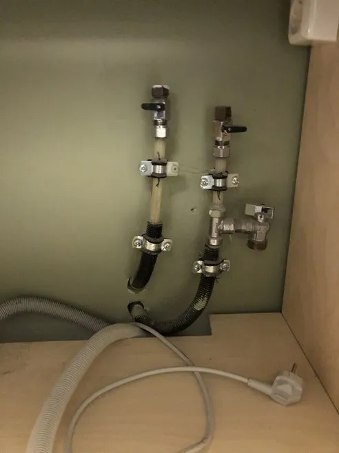 To stengekraner med slanger på veggen i et skap, med avløpsslange og støpsel under